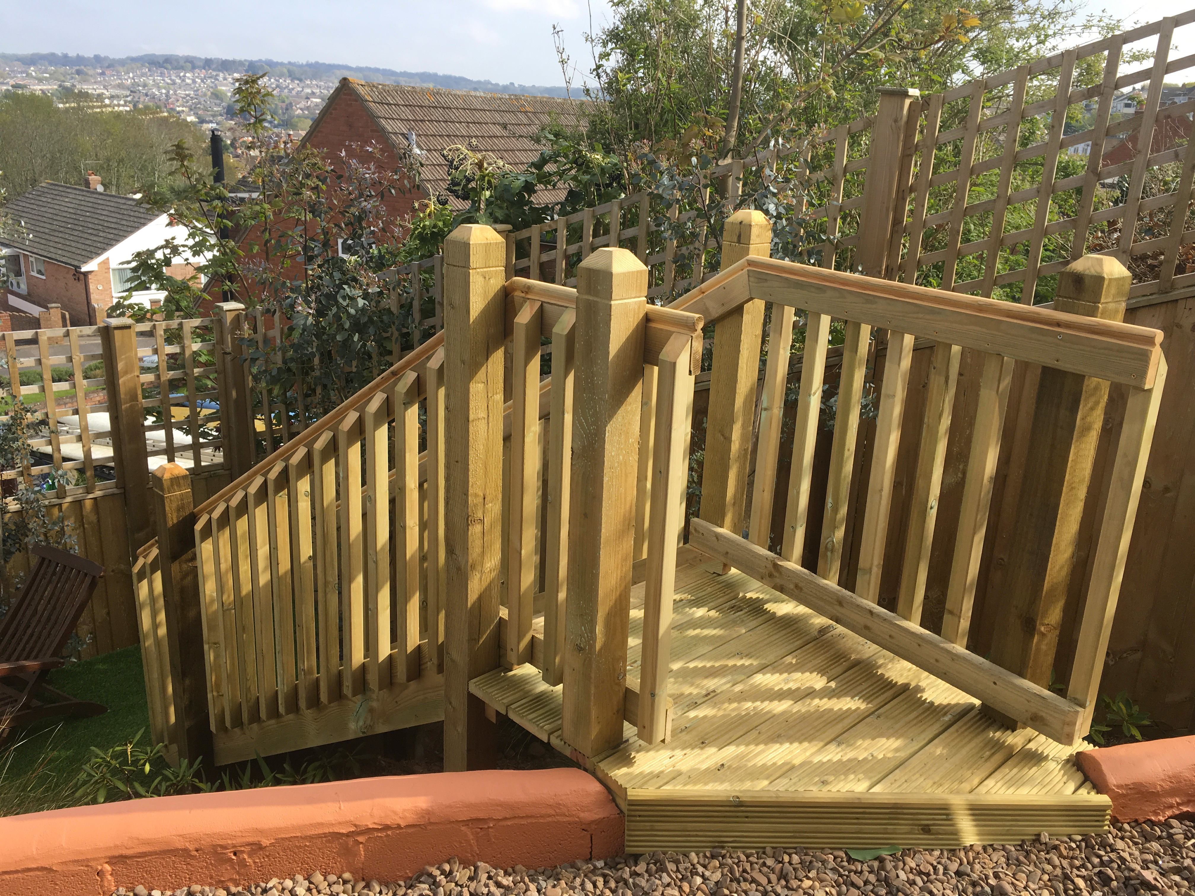 Wooden staircase in garden with railings and lattice fencing, offering a view of houses and landscape.