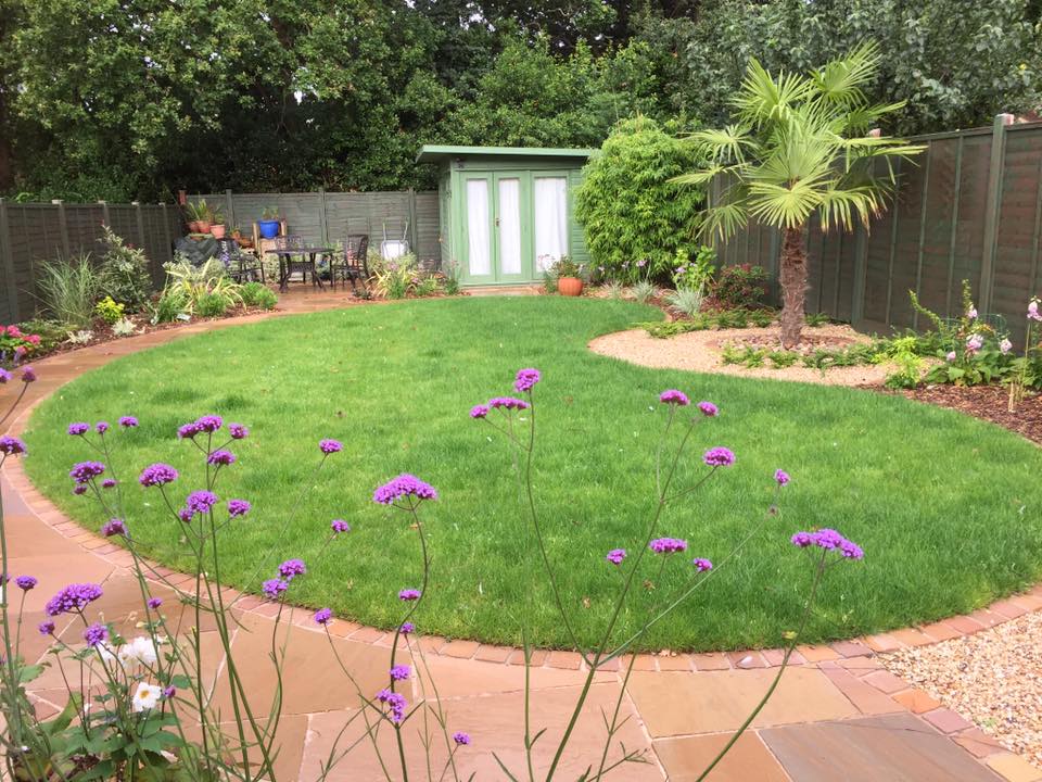 Well-maintained garden with circular lawn, purple flowers, patio, shed, and trees.